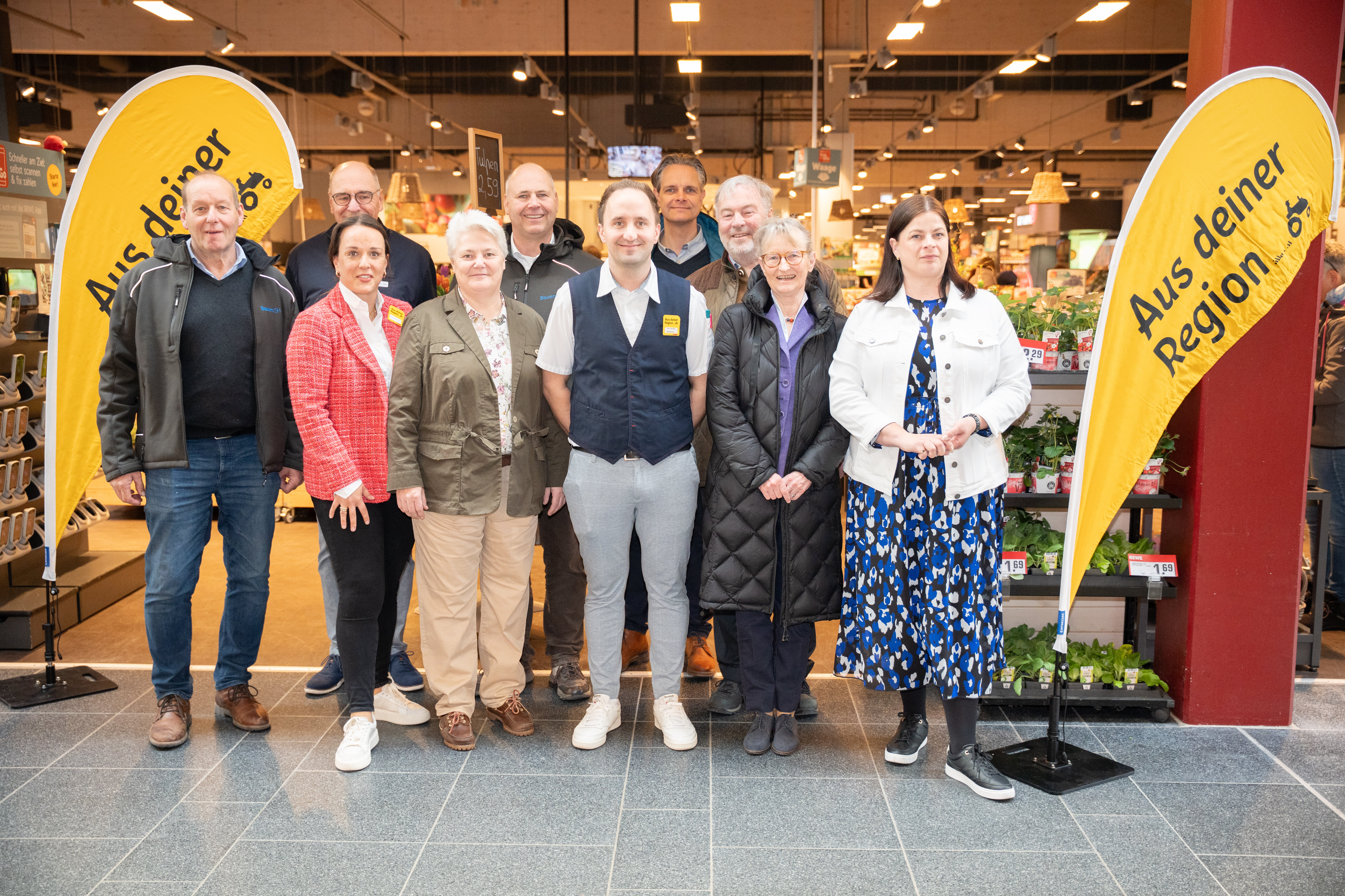 Eine Gruppe von zehn Leuten steht vor einem REWE-Markt und blickt in die Kamera.