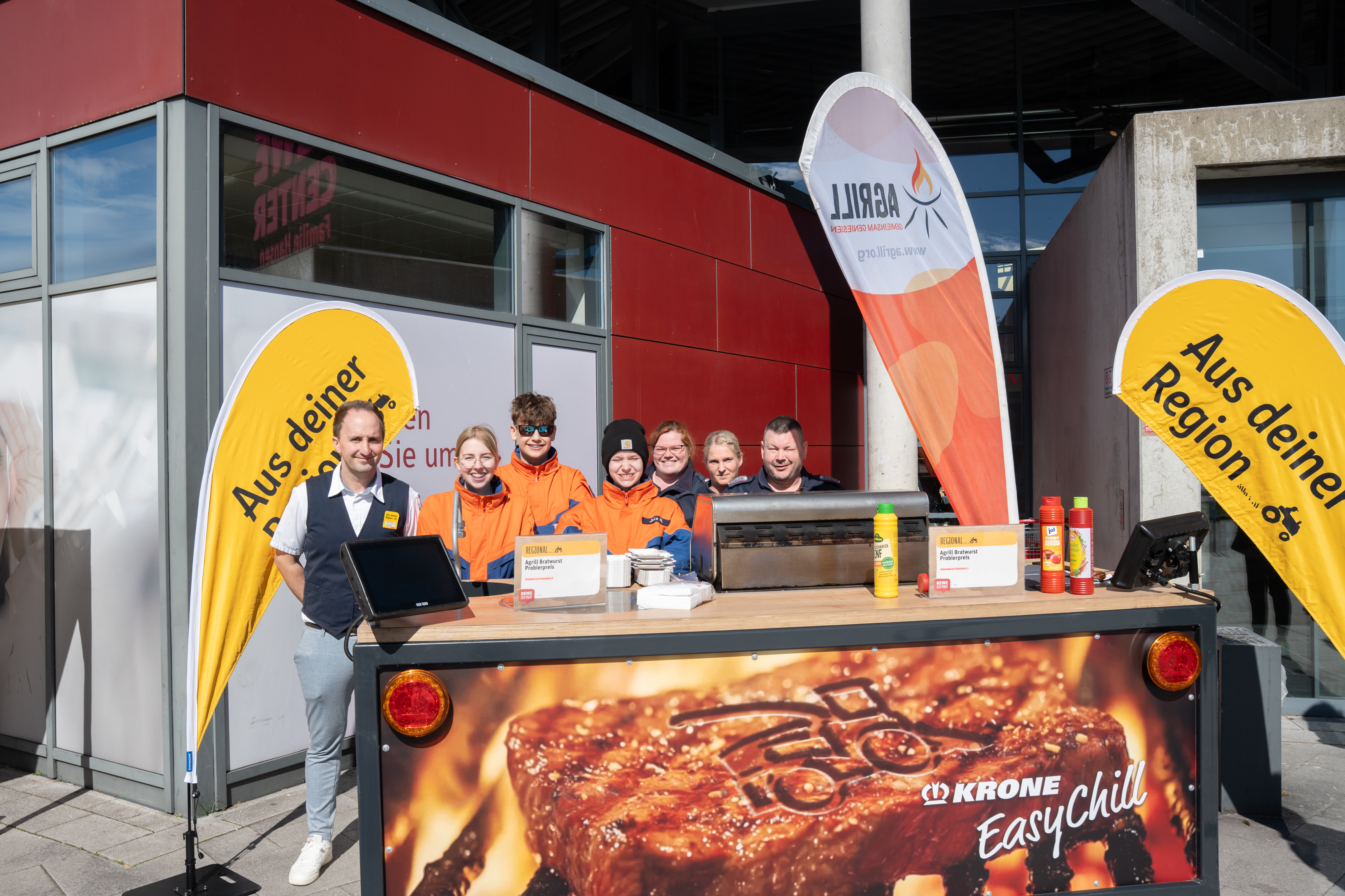 Mitglieder der Feuerwehr Büdelsdorf stehen hinter einem Grillstand und blicken gemeinsam mit dem REWE-Kaufmann in die Kamera. 