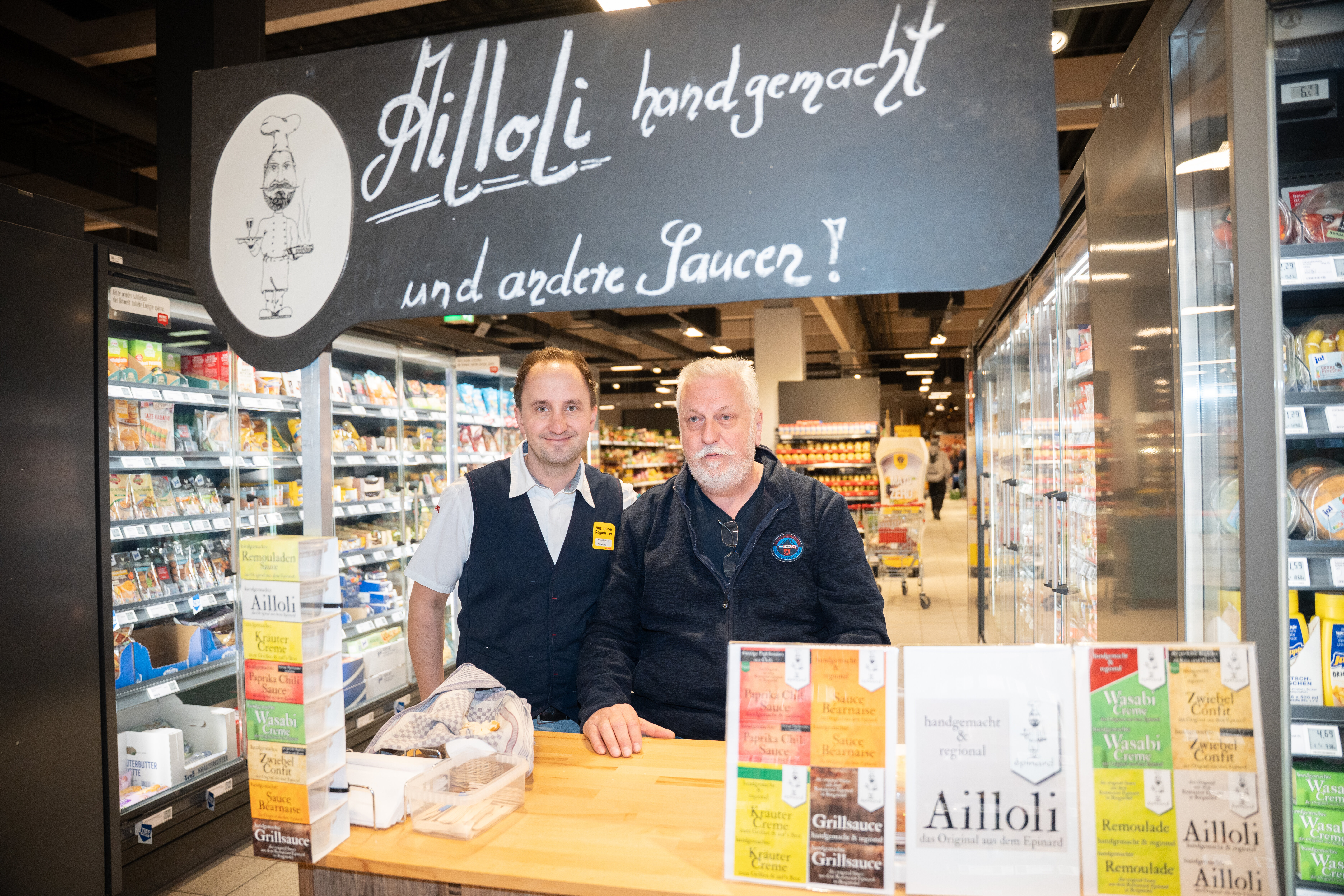 Der Kaufmann blickt gemeinsam mit einem Lieferanten in die Kamera, im Hintergrund sind Regale im Markt zu sehen.