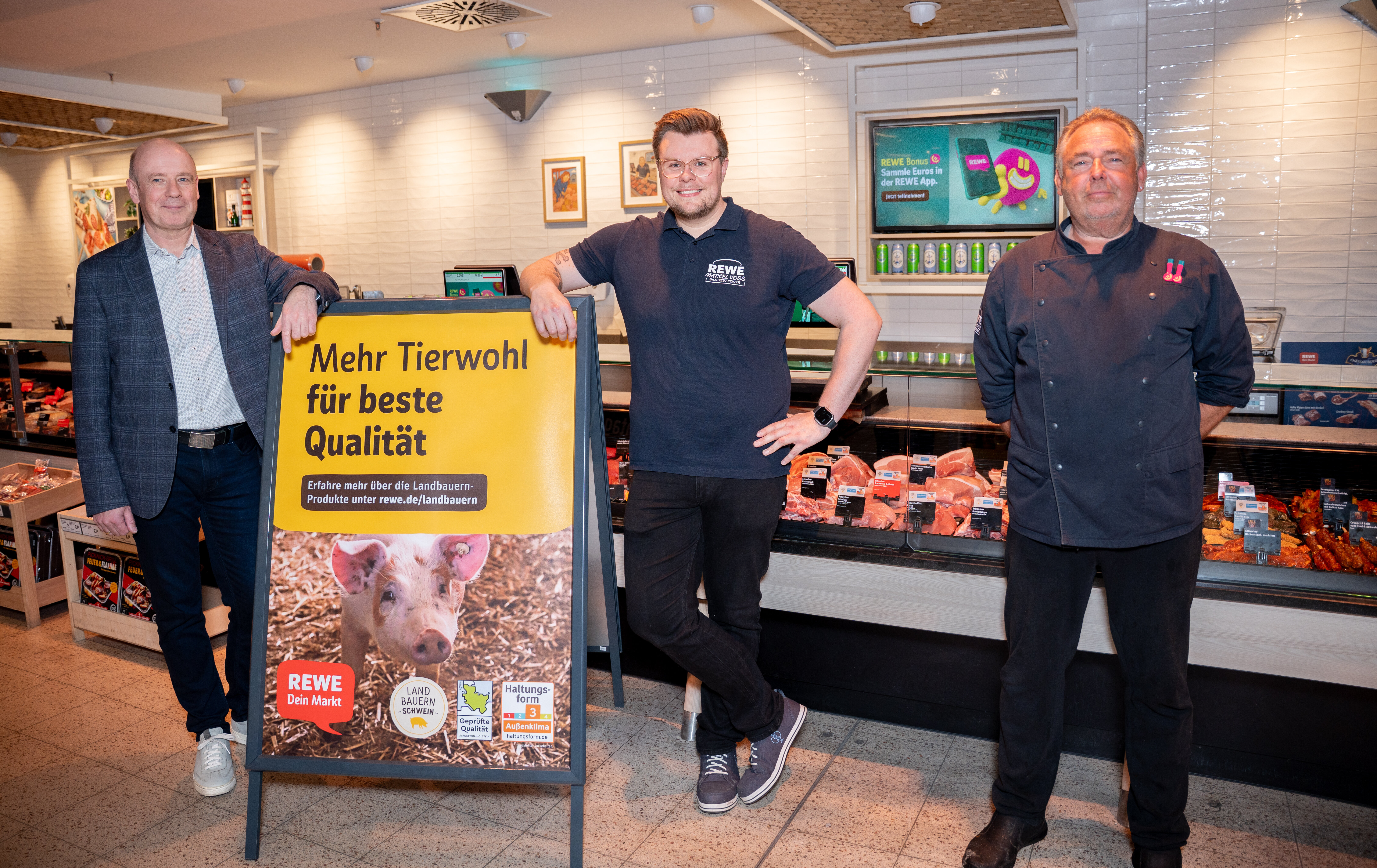Drei Personen posieren vor Frischetheke mit dem Werbeplakat zum neuen Schweinefleischprogramm.
