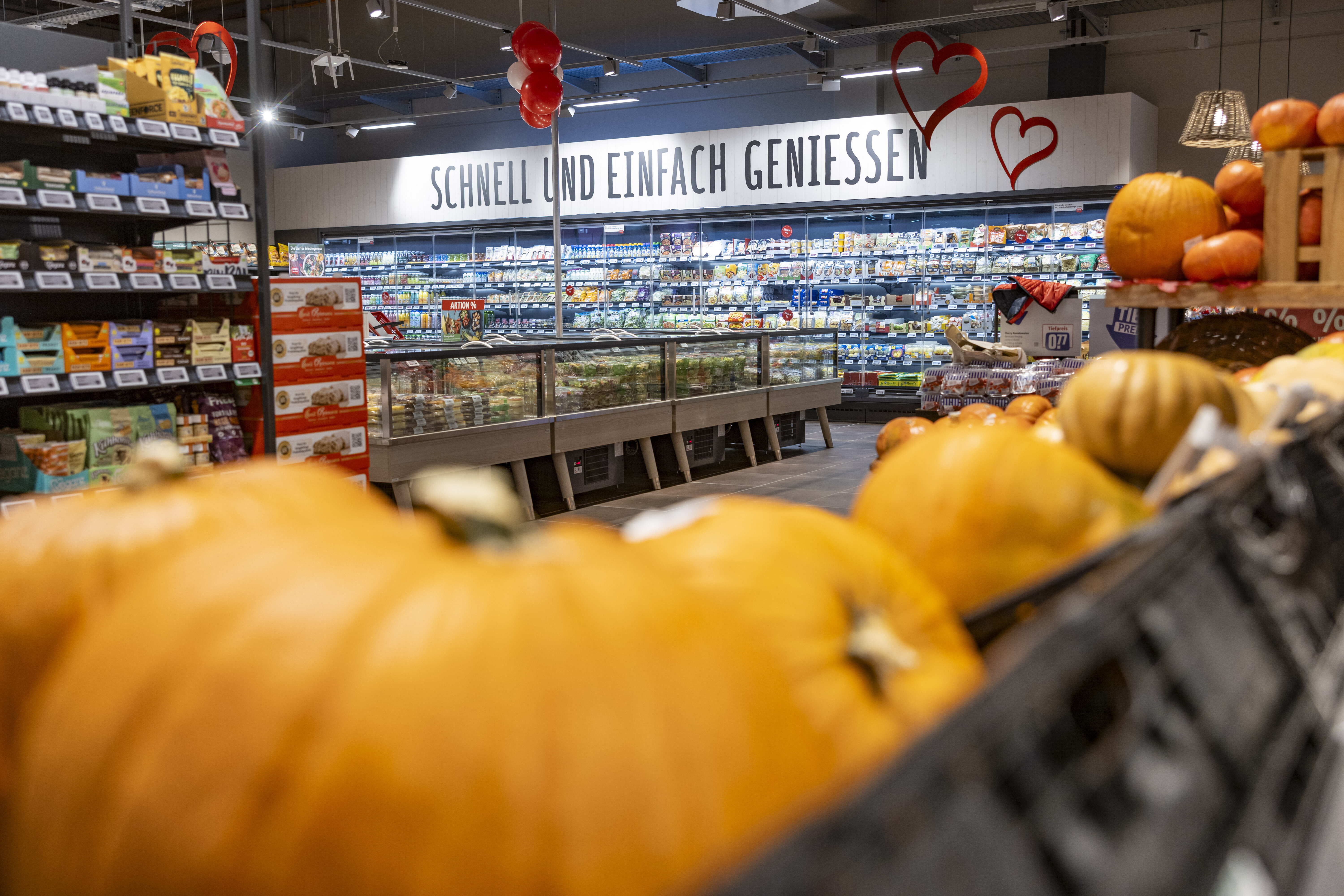 Innenansicht der Obst- und Gemüseabteilung mit Kürbissen unscharf im Vordergrund und Kühlregalen im Hintergrund mit der Aufschrift "Schnell und einfach genießen"
