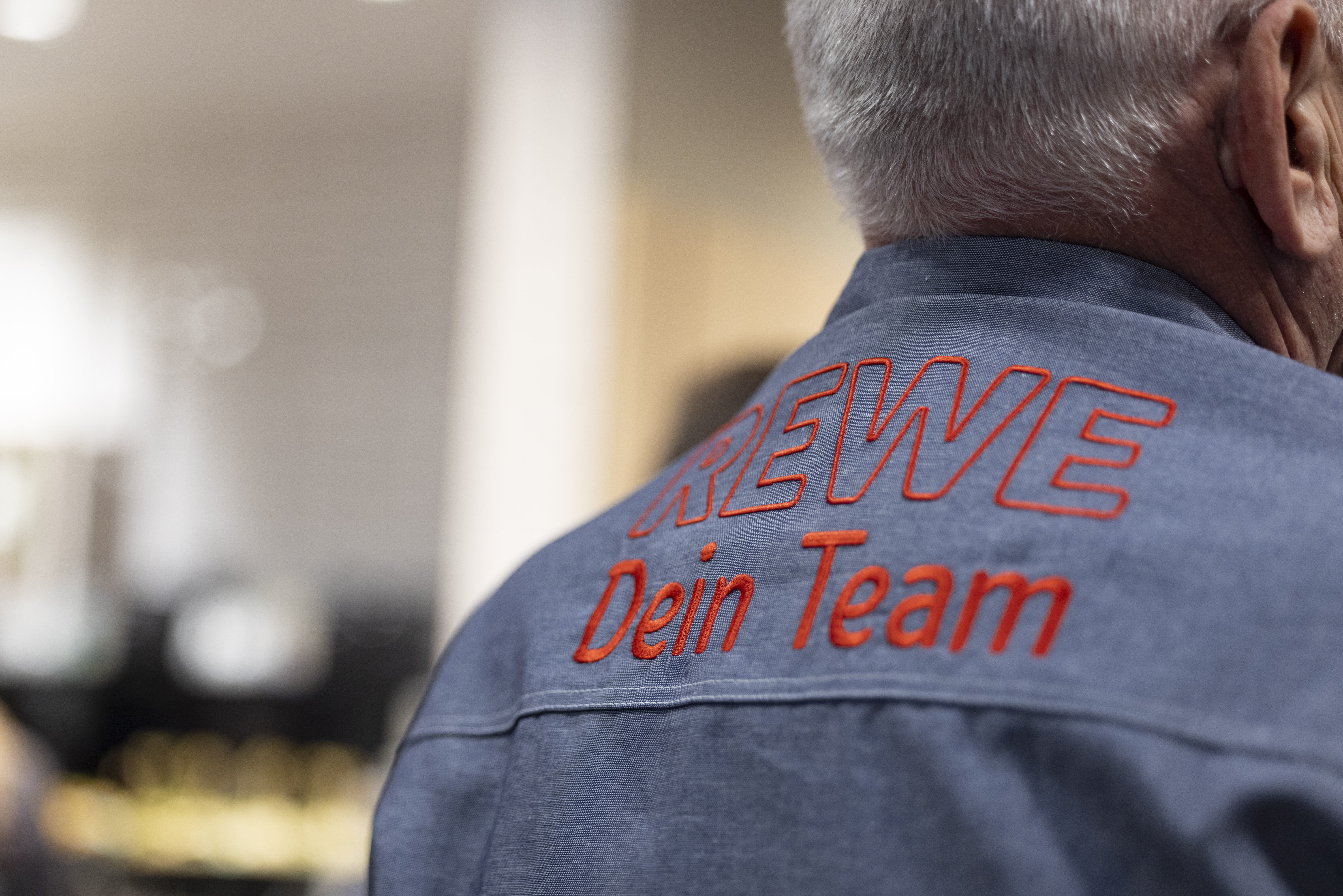Ein Mitarbeiter mit einem Kleidungsstück mit "REWE - Dein Team" als Aufschrift auf dem Rücken. Man sieht nur den Schulterbereich und den Hinterkopf