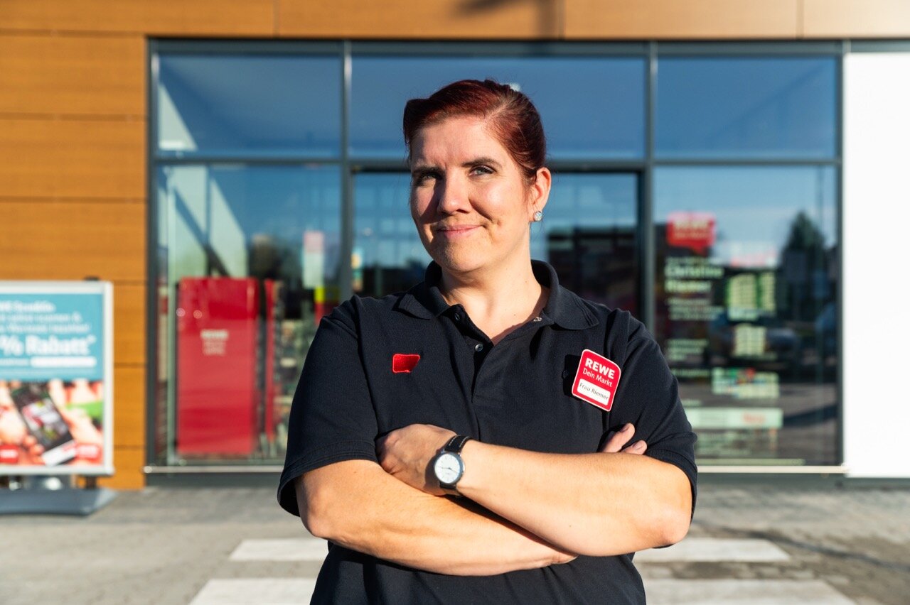 Kauffrau Christine Riemer steht vor ihrem REWE Markt und schaut in die Kamera. 