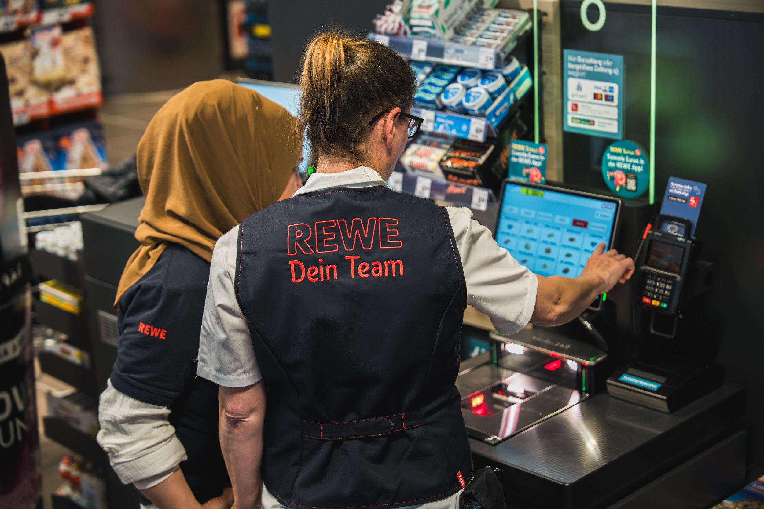 Zwei Mitarbeiterinnen an einer Self-Ceckout Kasse mit dem Rücken zur Kamera