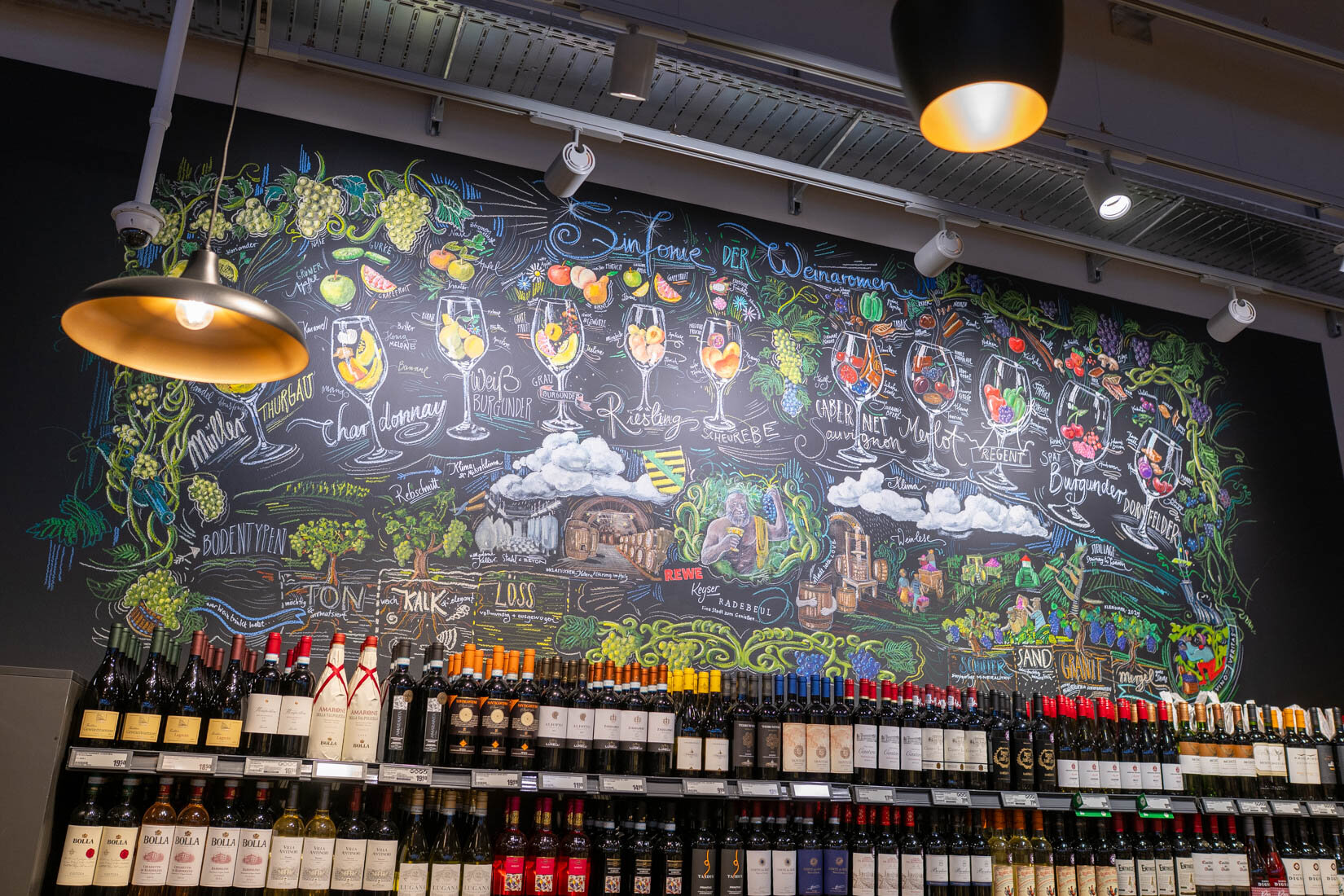 Weinflaschen stehen in Regalen, an der Wand hängt eine große Tafeln mit bunten Kreidezeichnungen.