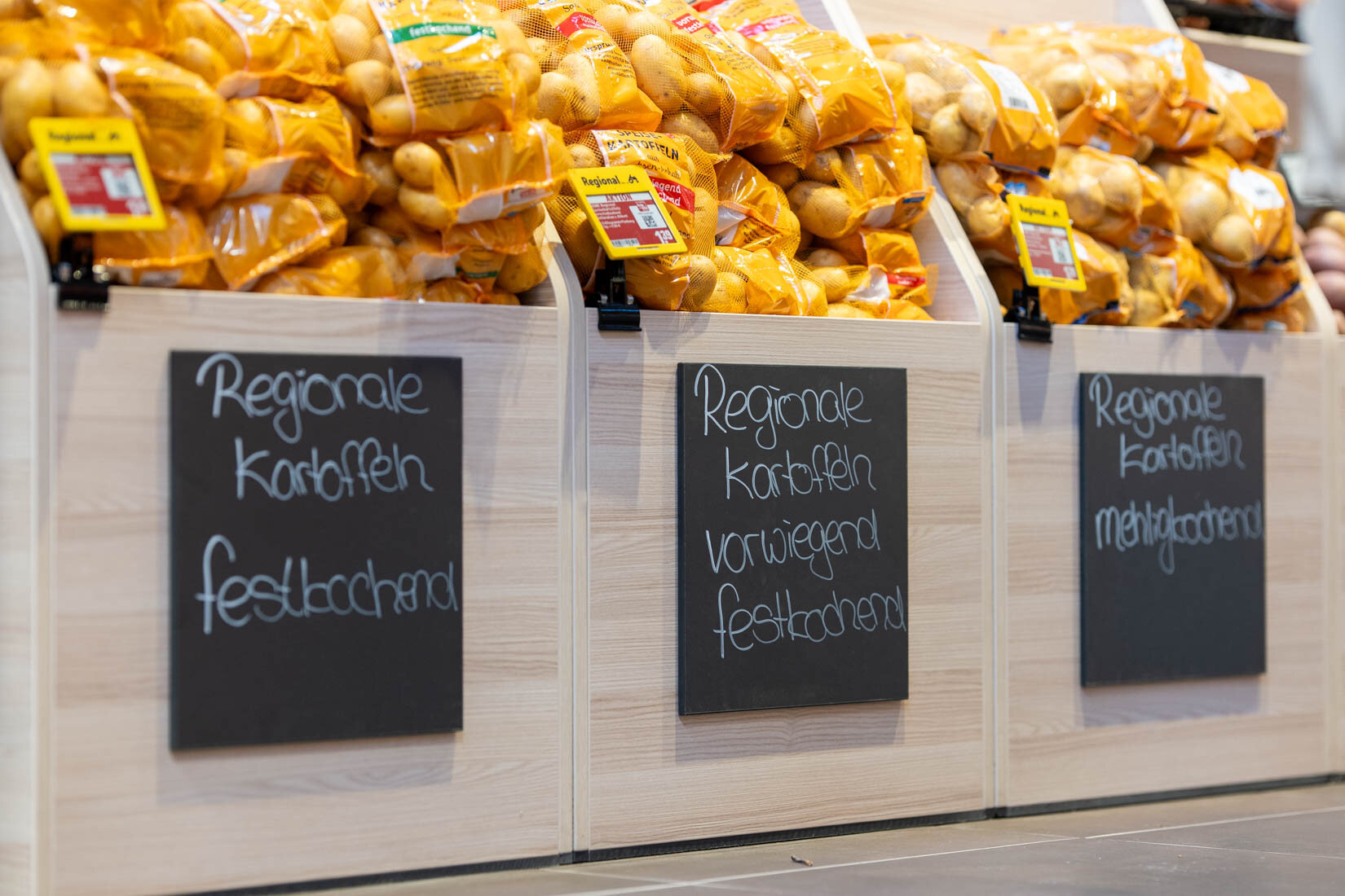 Kartoffeln im Netz liegen in Holzregalen, davor hängen Tafeln mit der Kreideaufschrift "Regionale Kartoffeln".