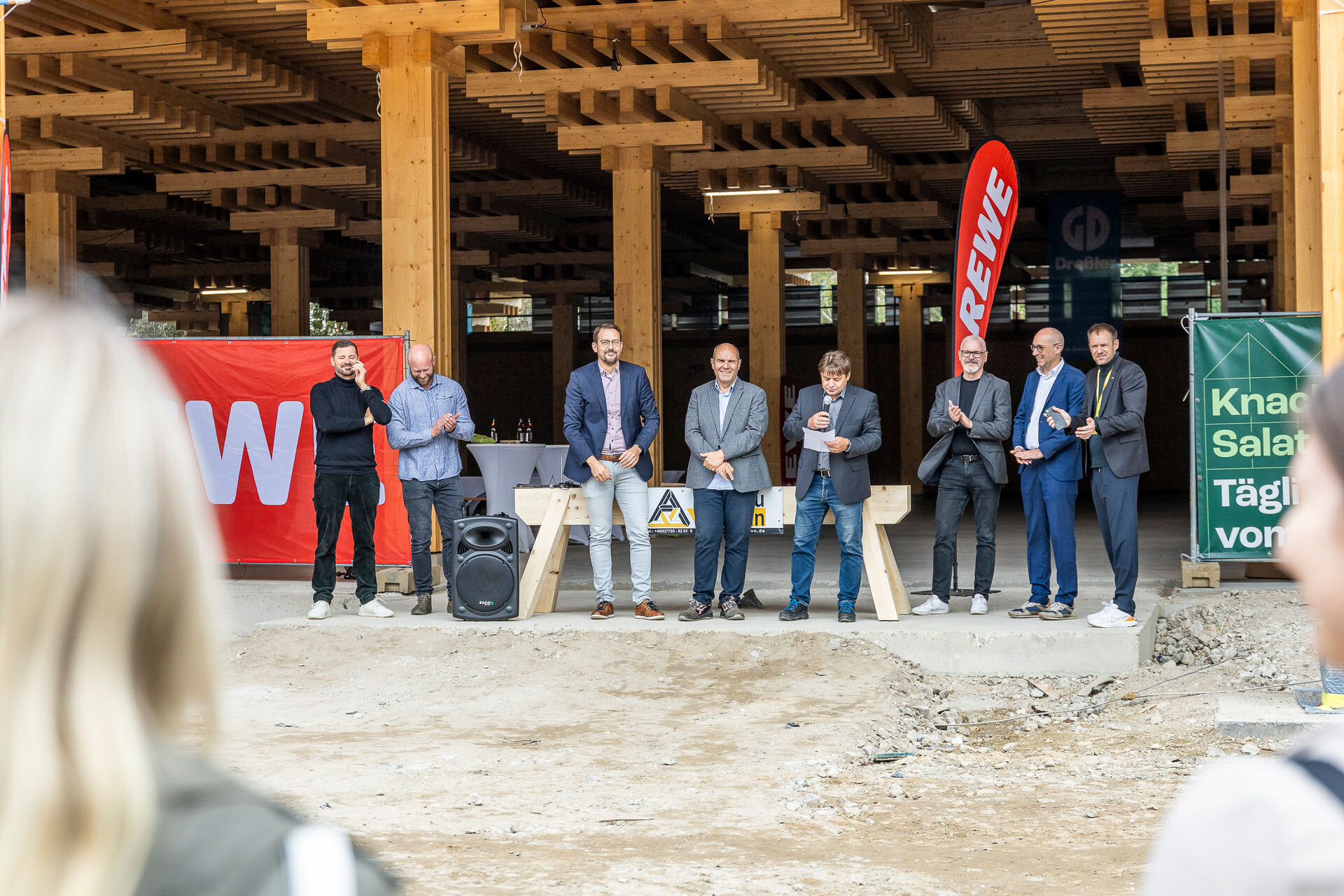 Sieben Männer vor Publikum vor einem Gebäude das sich im Bau befindet aus der Entfernung.