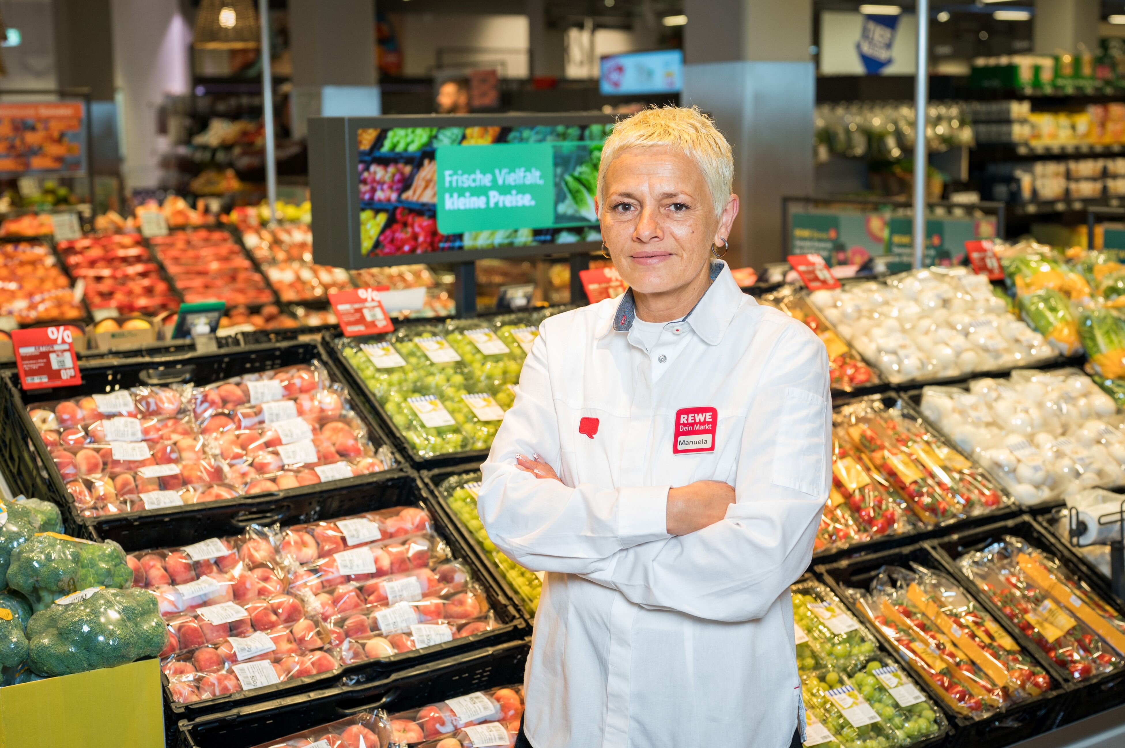 Eine Frau steht mir verschränkten Armen vor der Obst- und Gemüseabteilung eines Supermarktes.