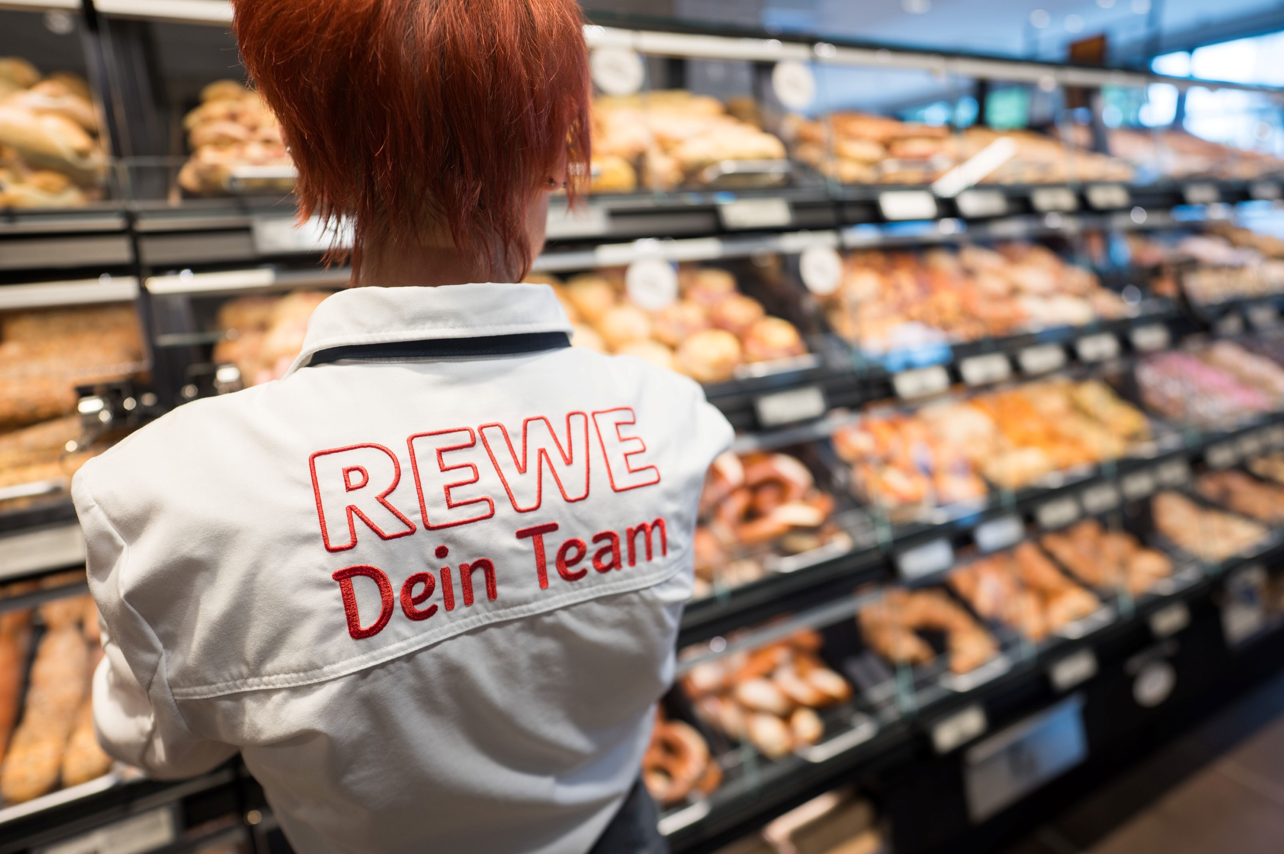 Eine Frau steht vor der REWE-Brotwelit mit dem Rücken zur Kamera. Auf dem Rücken ist der Schriftzug "REWE Dein Team" zu lesen.