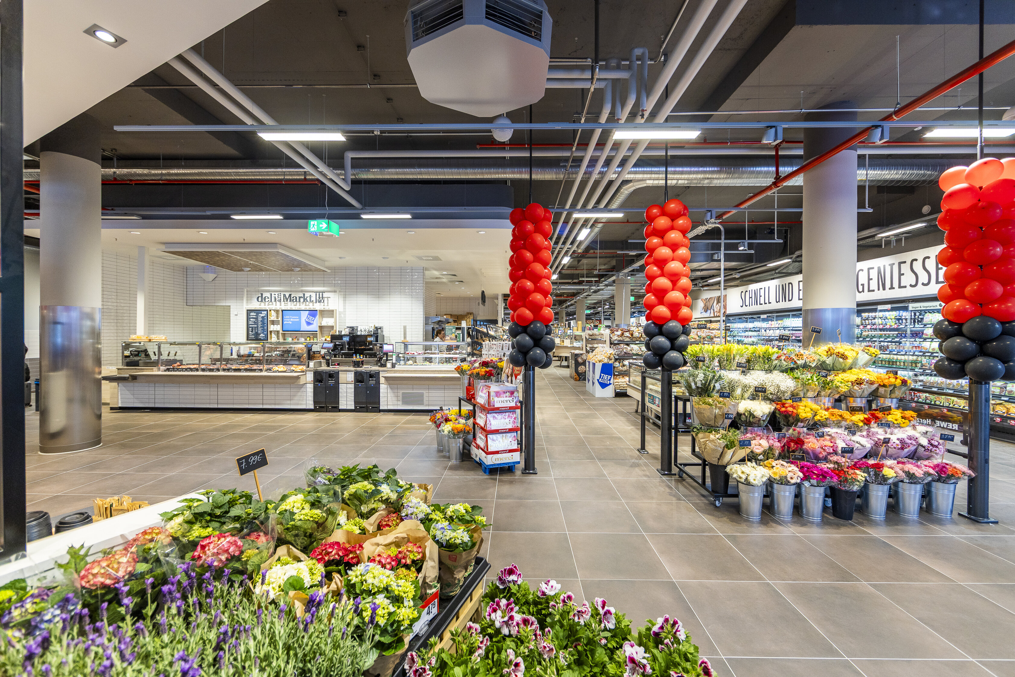 Der Eingangsbereich eines REWE Marktes. Link ist ein deli mit Speisen zu sehen. 