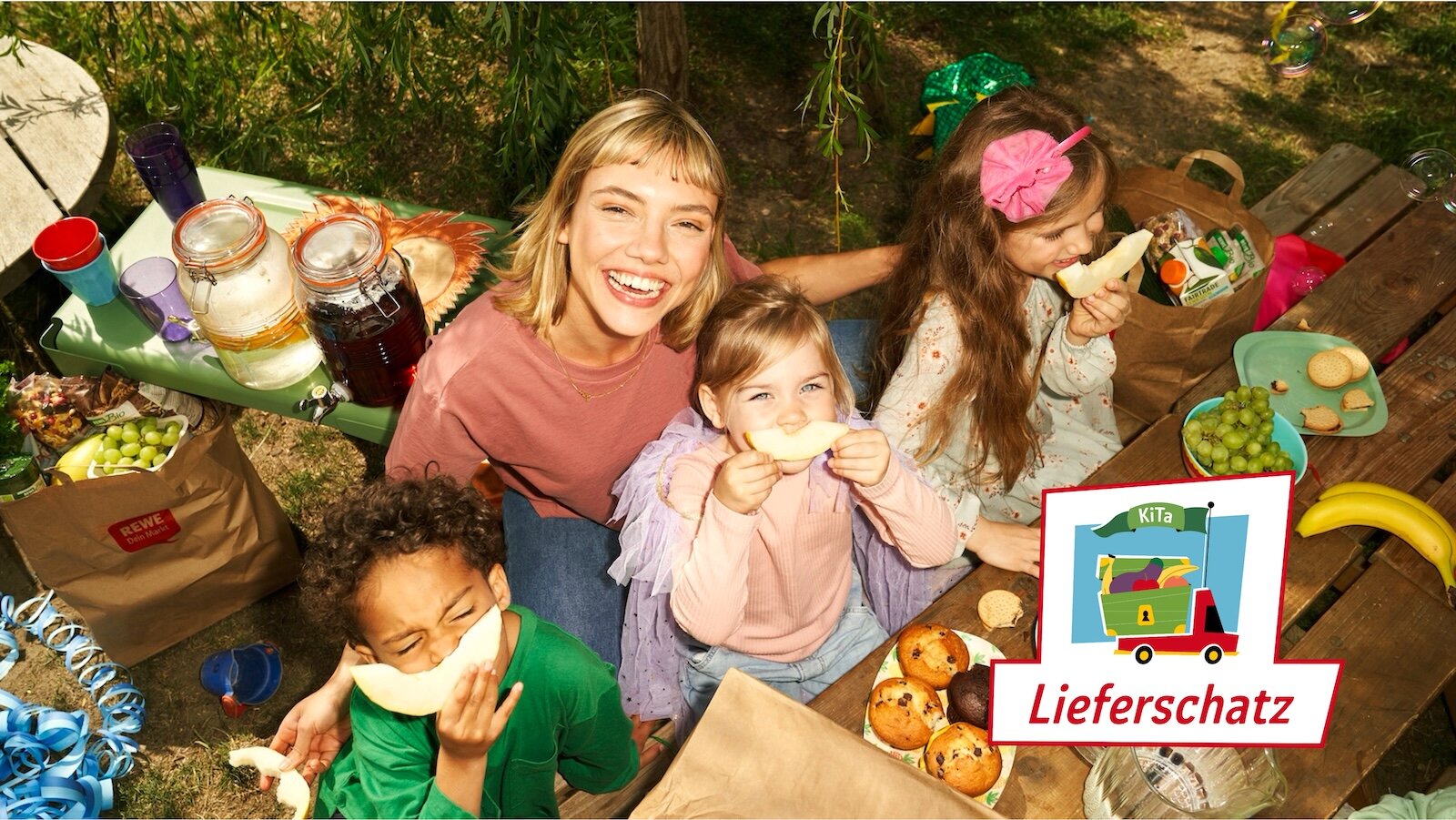 Drei Kinder und eine Erzieherin sitzen am Tisch im Park und essen Melone.