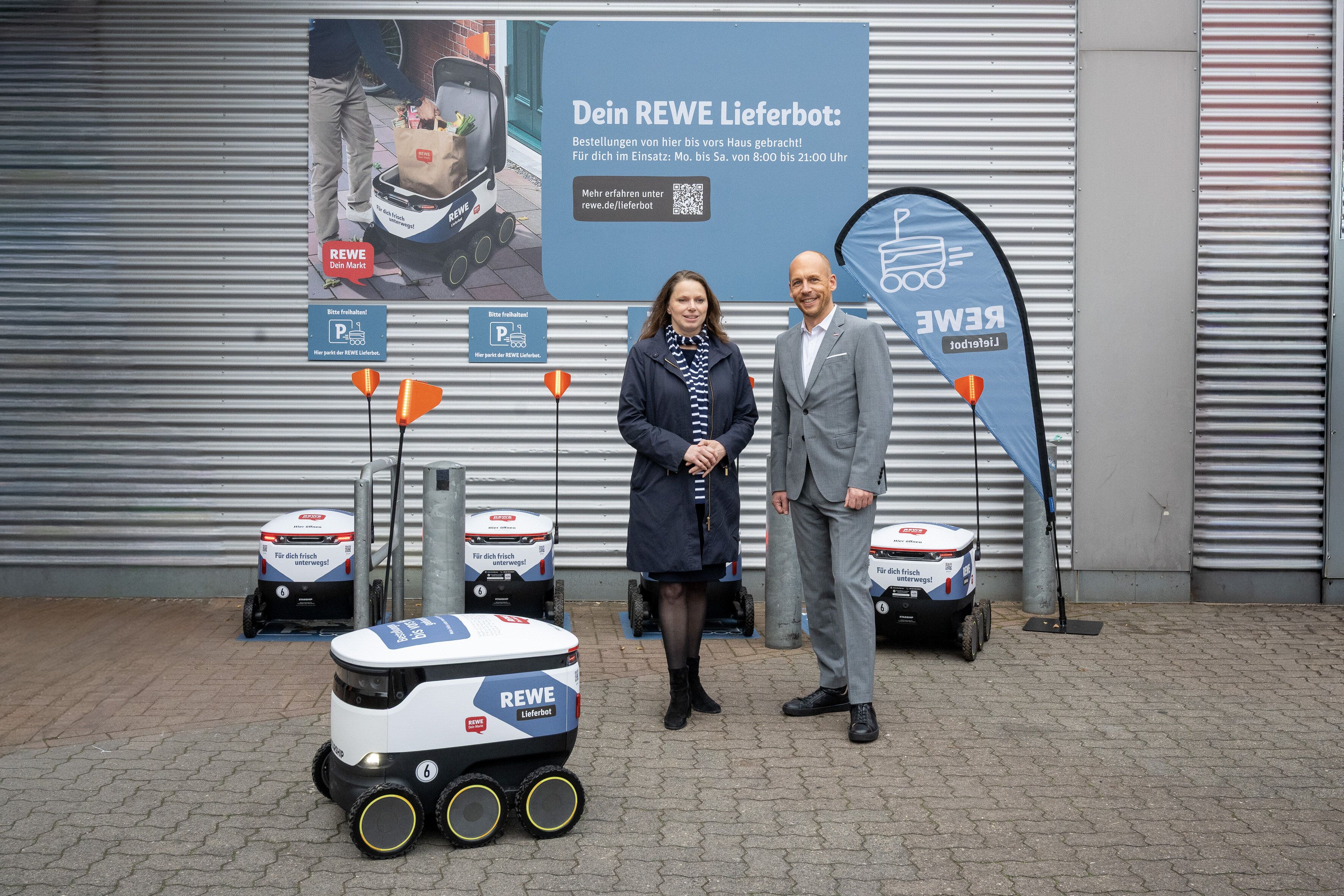 Zwei Personen stehen vor grauer Wand mit Lieferrobotern im Hintergrund.